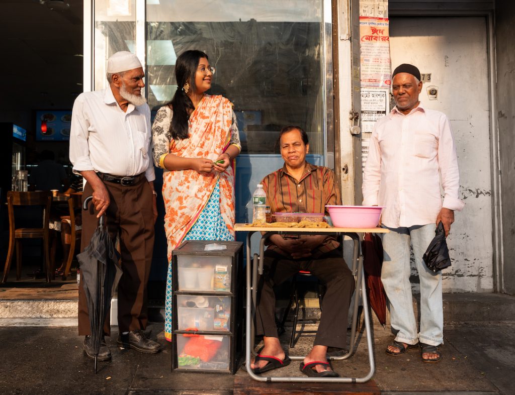 4 people gathering in front of a Brooklyn shop, one seated with a tray holding food.